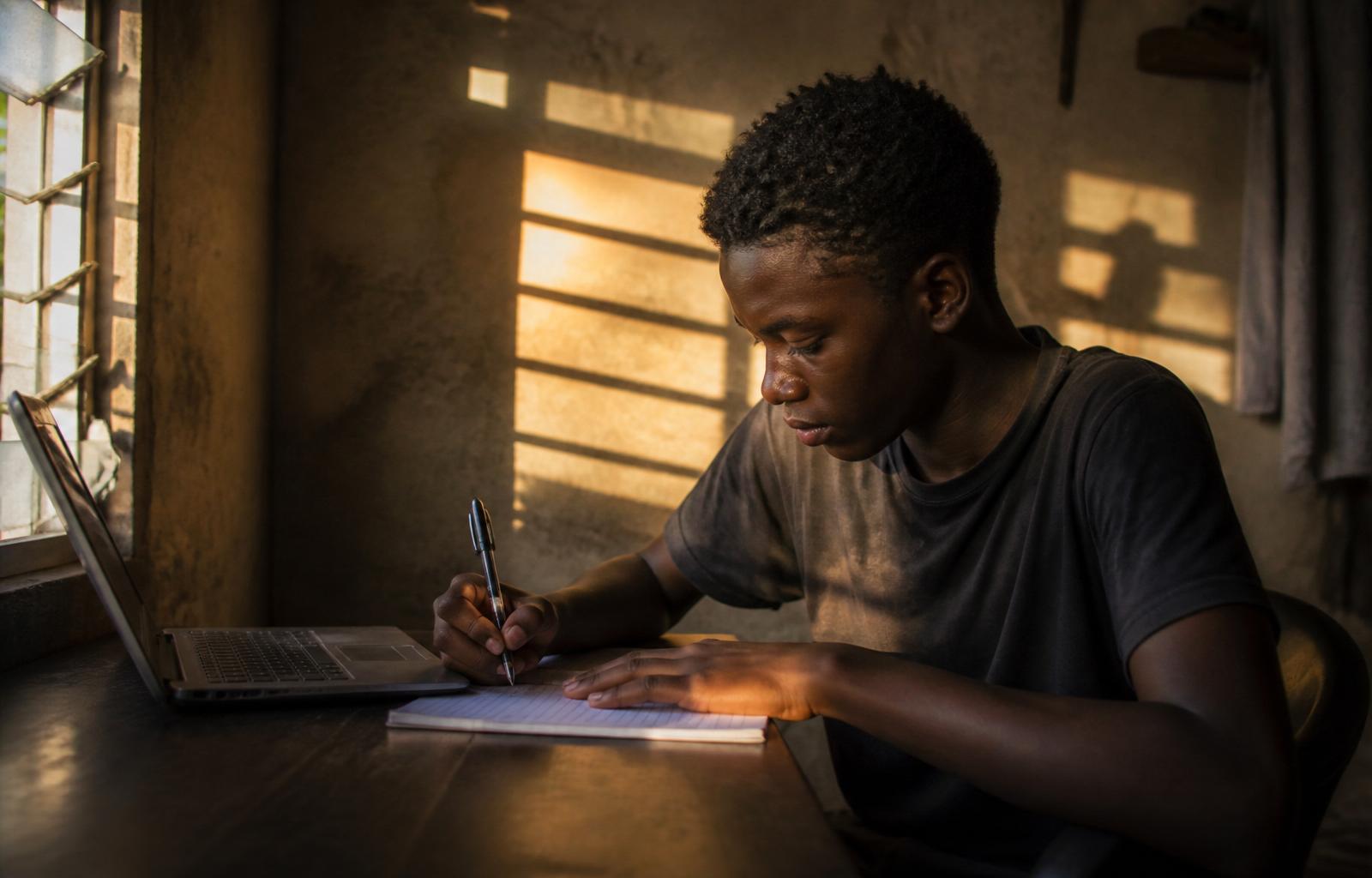 Young Jamaican student focused on a laptop showing code and AI dashboards — driven by purpose